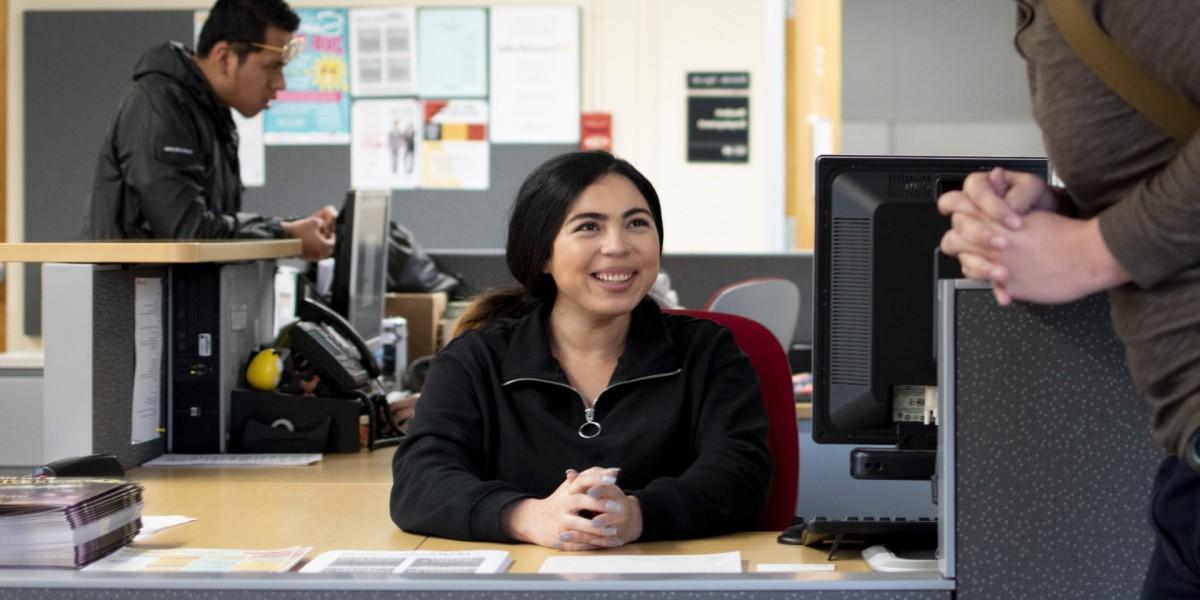 Student employee sits at office desk, smiling at a person standing at the counter.