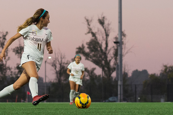 Image of a member of the  women's soccer team on the pitch at dusk, pink sky behind her. 