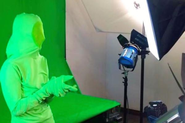 Image of a student in a green suit in front of a green screen being filmed. 