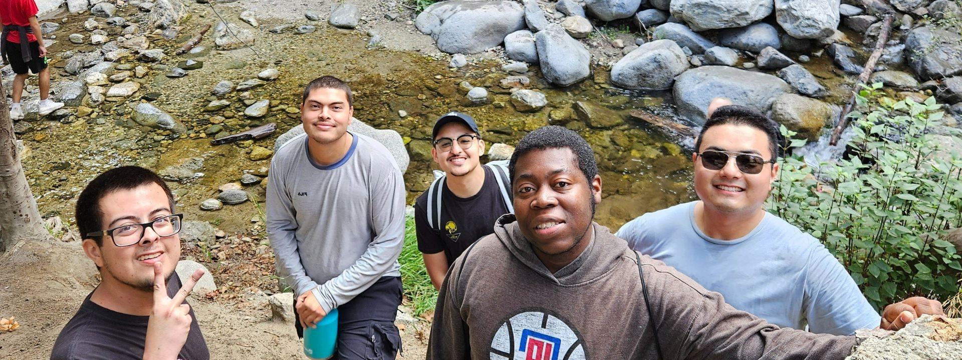 5 Men smiling in front of a stream of water and boulders.