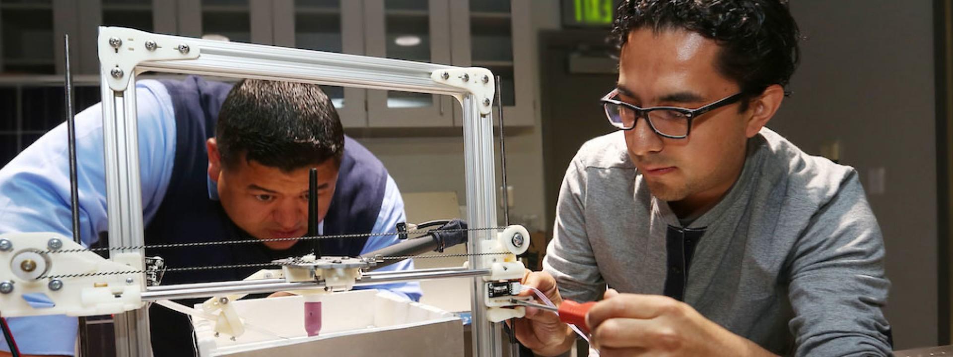 Image of two engineering students looking at a metal frame device intently. 