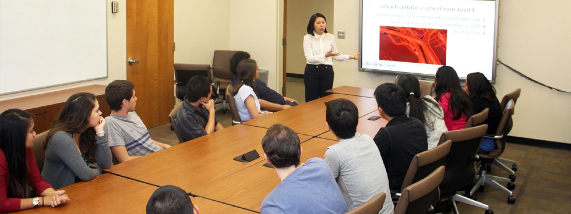 Group of Cal State LA students in a classroom around a conference table facing a projector screen as a lecturer teaches.